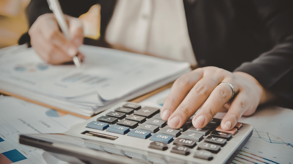 Nahaufnahme einer Person, die an einem Schreibtisch sitzt und Finanzdokumente durchgeht. Die linke Hand hält einen Stift und bearbeitet ein Diagramm, während die rechte Hand einen Taschenrechner benutzt. Der Hintergrund zeigt verschwommene Papiere und Diagramme.